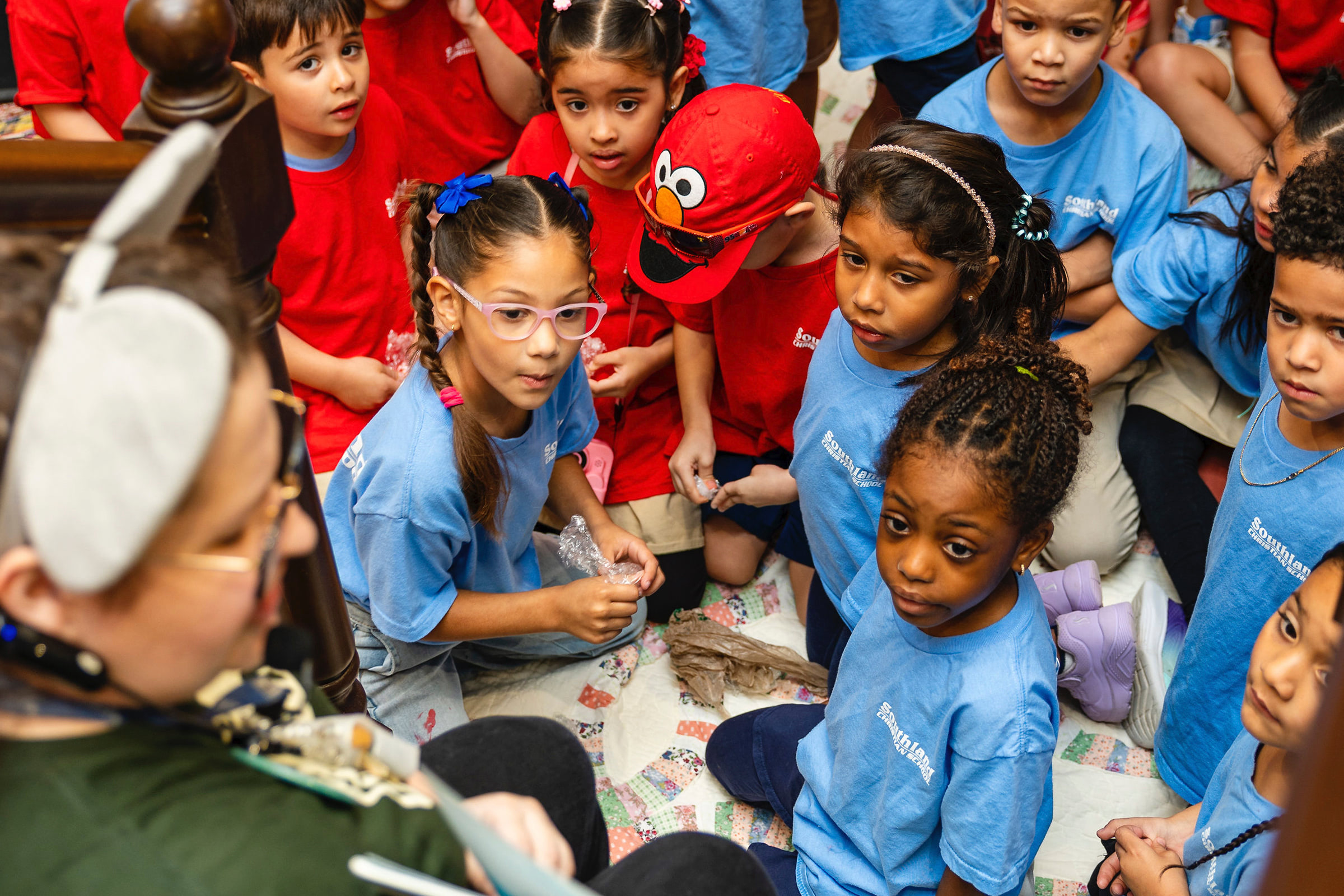 A group of kids paying attention to a volunteer in elephant ears, reading a children's book